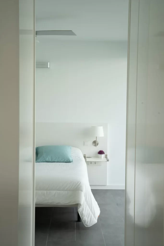 Dormitorio blanco visto entre puertas correderas, con cama de cojín turquesa y aplique junto al cabecero