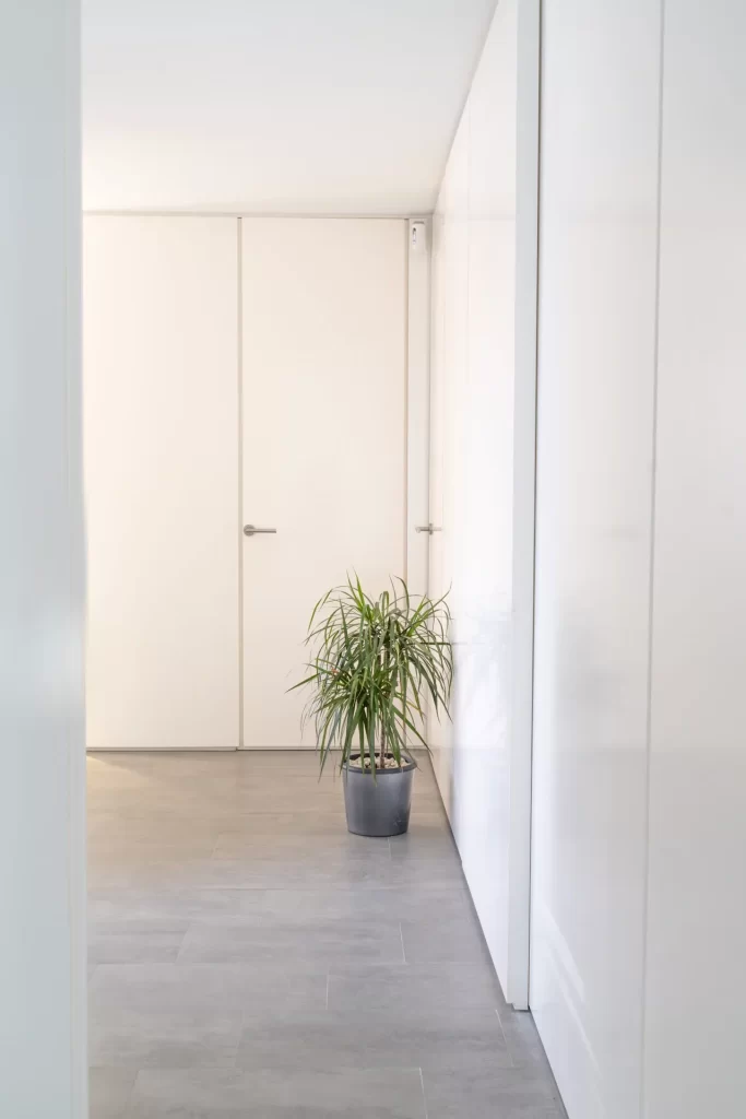 Pasillo blanco con puertas enrasadas, suelo gris de baldosa y una planta en maceta junto a la pared