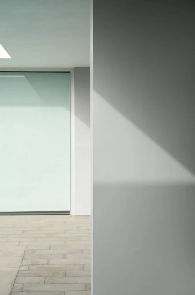 Haz de luz diagonal sobre un muro gris junto a un gran ventanal de vidrio y pavimento de piedra