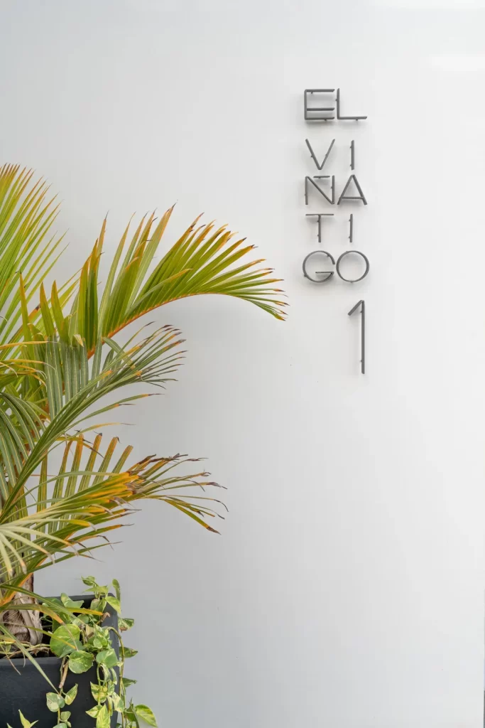 Letras metálicas en vertical sobre una pared blanca junto a una palmera en jardinera negra con potos