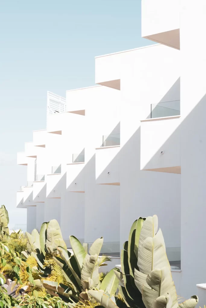 Fachada blanca escalonada con balcones en voladizo y barandillas de vidrio sobre una hilera de plataneras