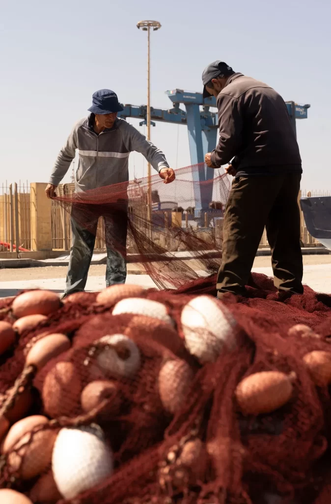 Dos pescadores extienden una red roja con boyas en un muelle con grúa azul