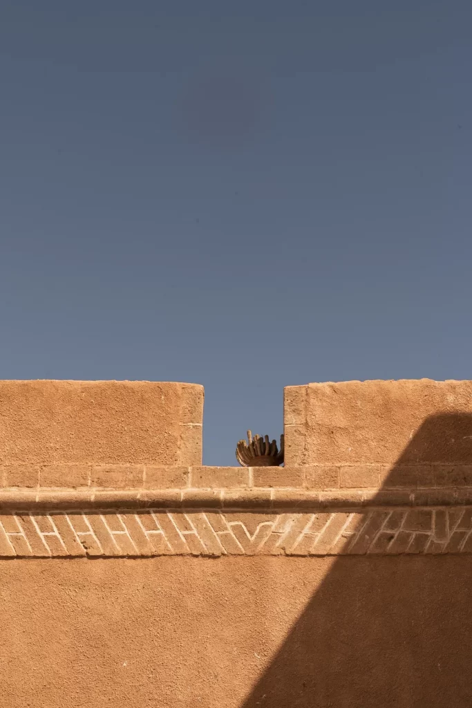 Muro ocre con almenas y relieve geométrico bajo un cielo azul, con un cactus asomando entre los merlones