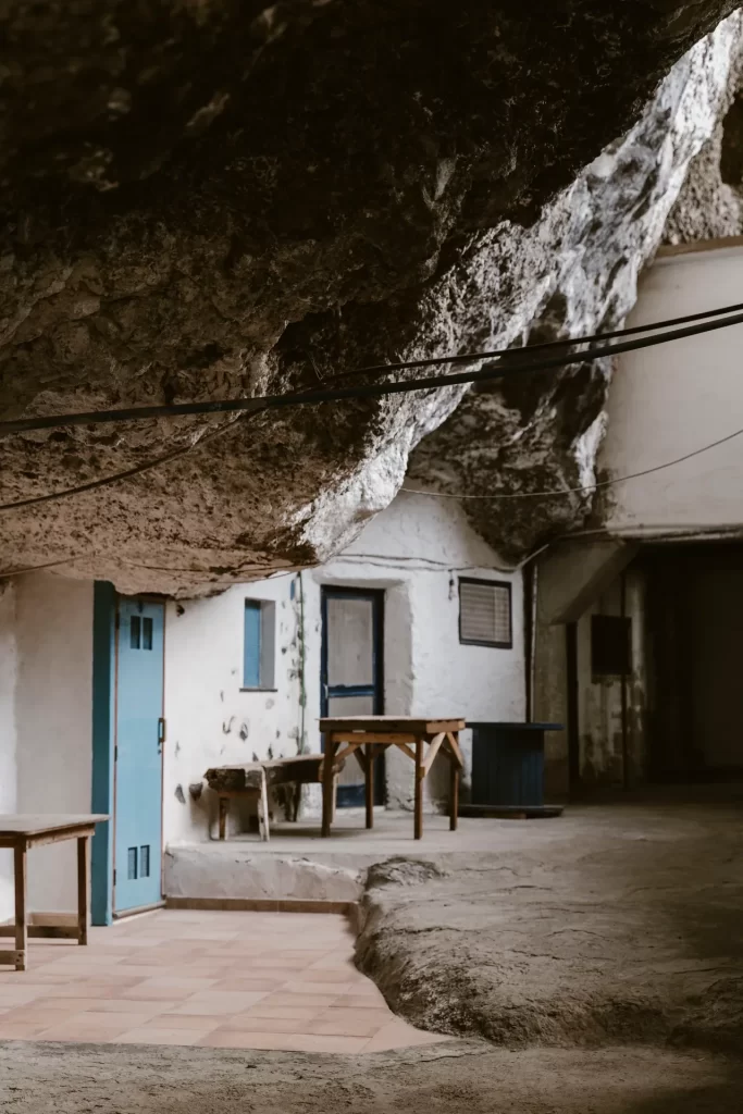 Casas encaladas con puertas azules y mesas de madera bajo el techo de roca de una gran cueva