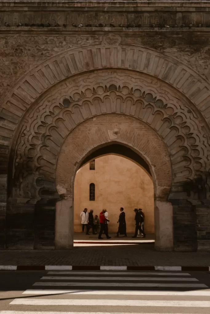 Personas caminando bajo un gran arco de piedra decorado, con paso de cebra delante