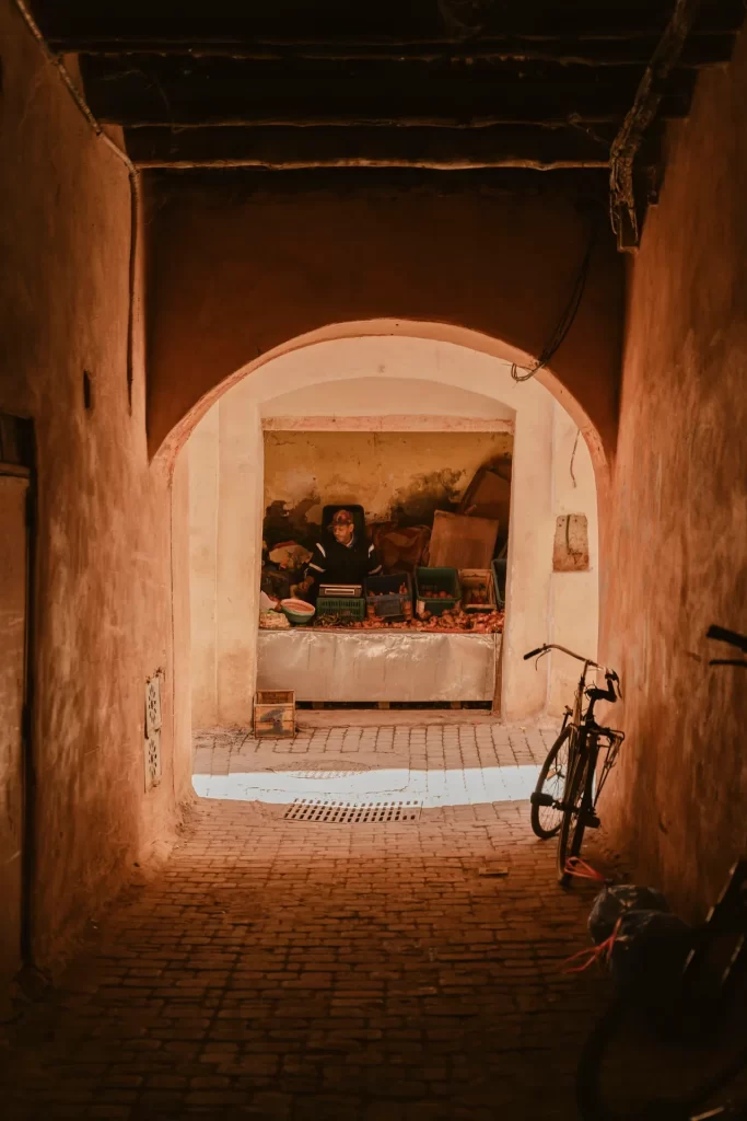 Pasaje cubierto de muros ocres que se abre en un arco hacia un puesto de fruta, con una bicicleta apoyada