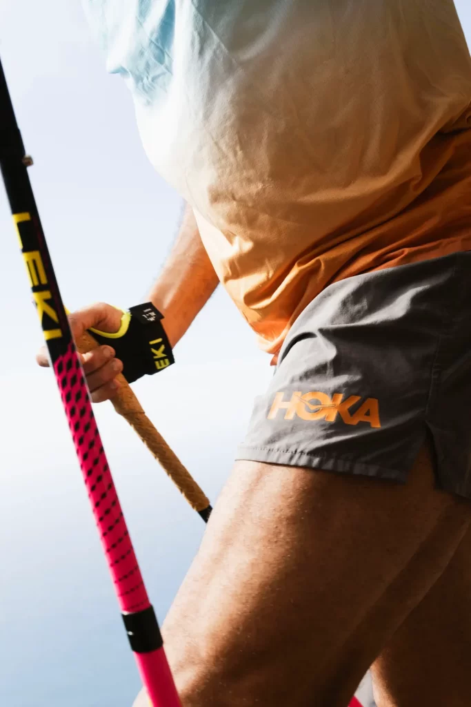 Detalle de corredor con pantalón corto Hoka sujetando bastones de trekking