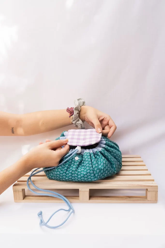 Manos abriendo un neceser de tela verde estampada con solapa de cuadros lilas sobre un palé de madera