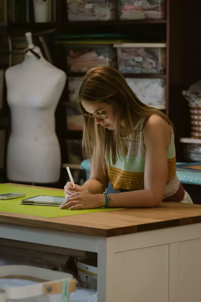 Mujer con gafas dibujando en una tableta sobre la mesa de un taller de costura con maniquí al fondo