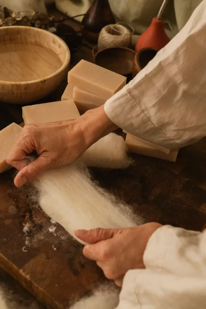 Manos extendiendo lana blanca sobre una tabla de madera rodeada de pastillas de jabón artesanal