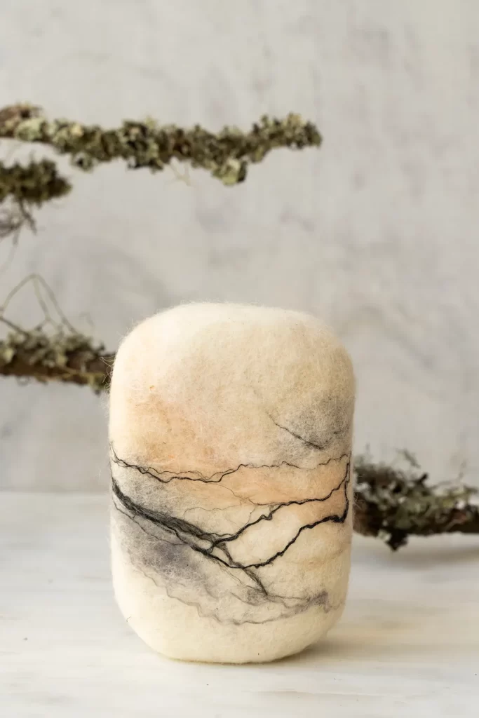 Jabón de fieltro con vetas negras y melocotón en pie sobre mármol blanco con ramas de liquen detrás