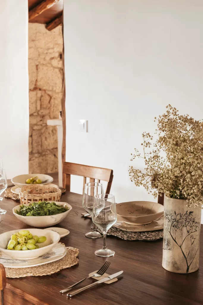Mesa de madera puesta con cuencos de cerámica, uvas, ensalada, copas y un jarrón con flores secas