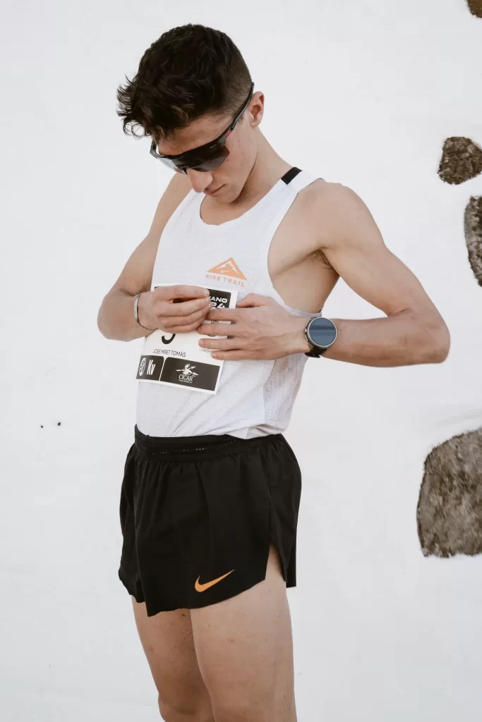Corredor con gafas de sol se ajusta el dorsal sobre la camiseta blanca ante una pared encalada