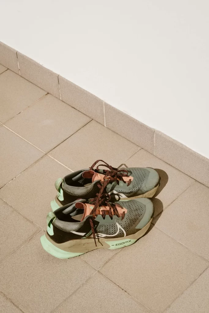 Zapatillas de trail usadas con cordones naranjas sobre un suelo de baldosas junto a una pared blanca