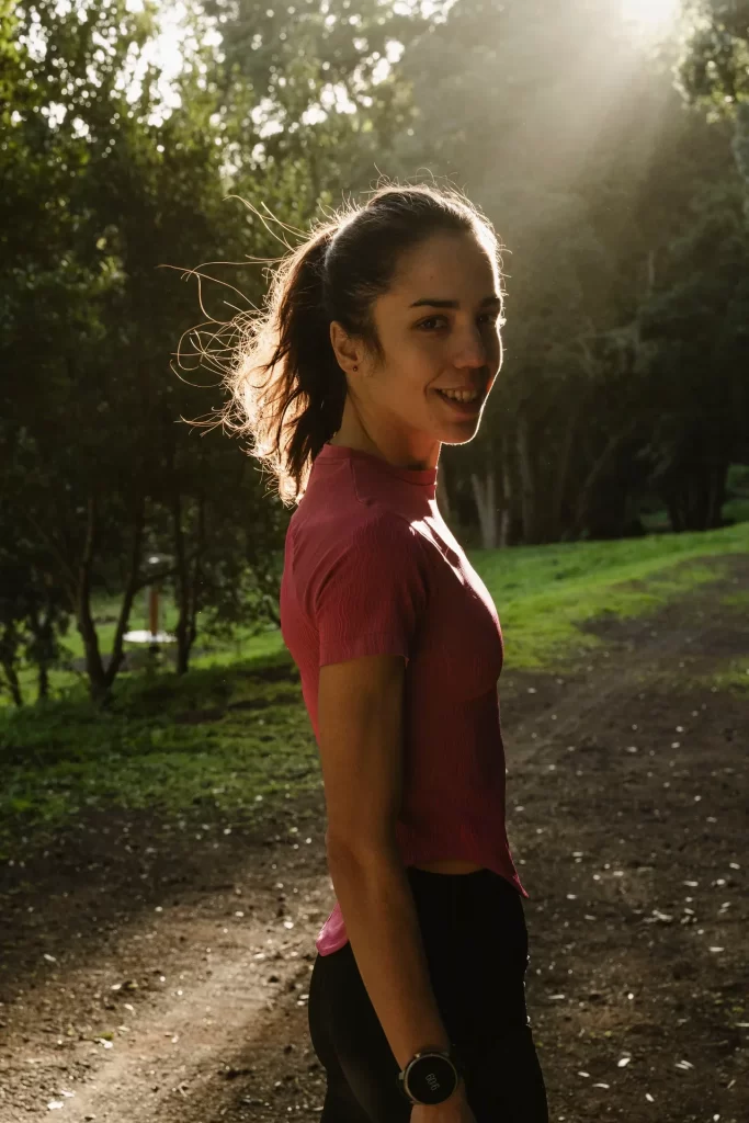 Corredora con camiseta rosa y coleta mira sonriente a cámara en un sendero del bosque a contraluz