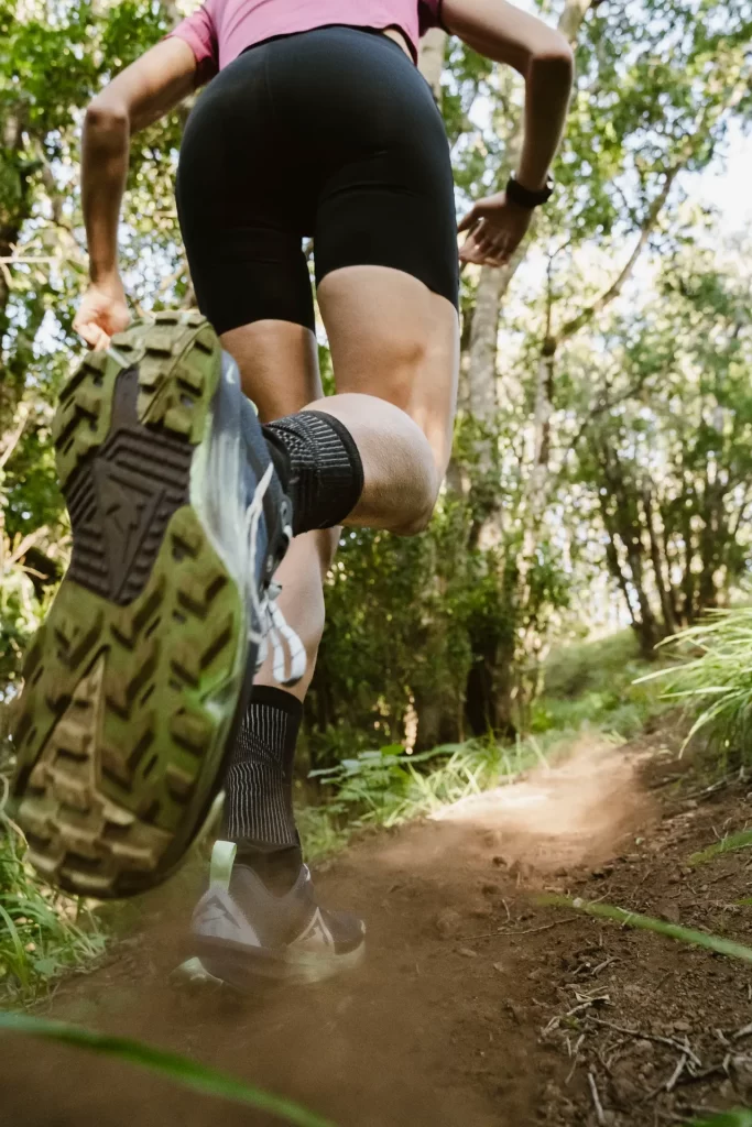 Suela de una zapatilla de trail en primer plano mientras una corredora sube un sendero entre árboles