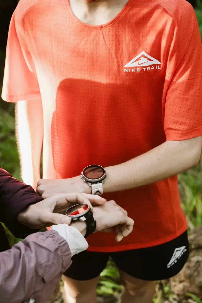 Corredores con camiseta naranja de trail comparan sus relojes deportivos en mitad del bosque