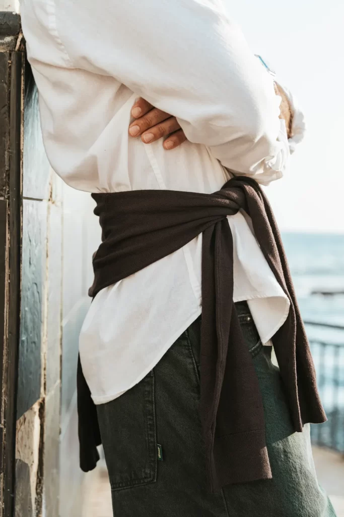 Detalle de camisa blanca con jersey marrón anudado a la cintura junto a una barandilla frente al mar