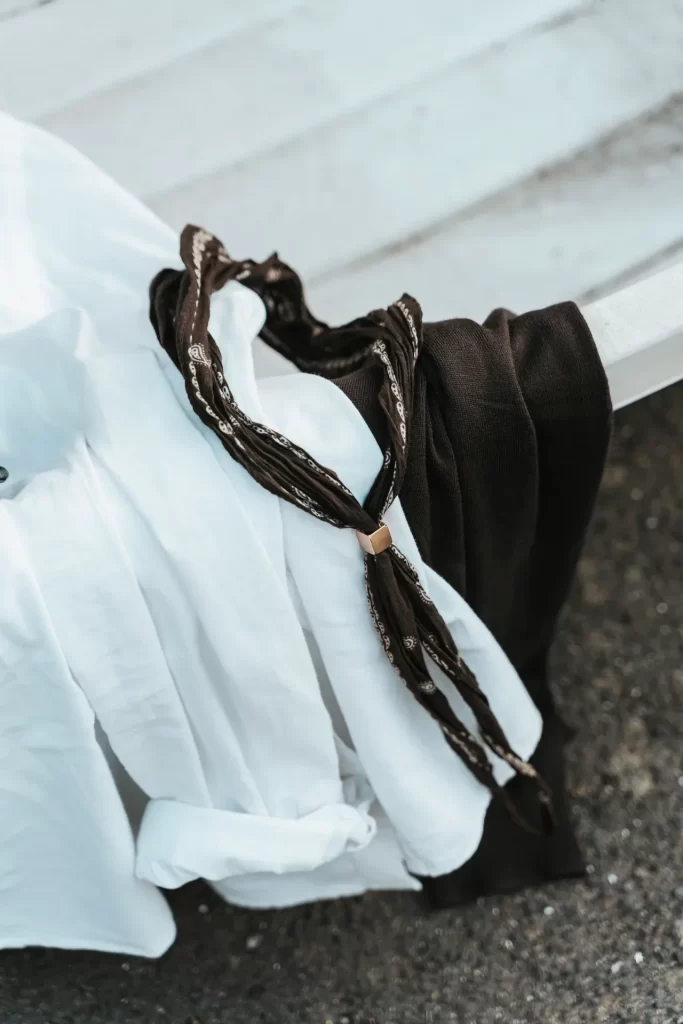 Camisa blanca, jersey marrón y pañuelo estampado con pasador dorado apoyados sobre un banco blanco