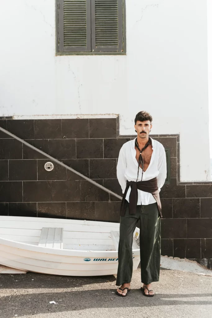 Hombre con camisa blanca y pantalón verde de pie junto a una barca blanca ante una fachada encalada