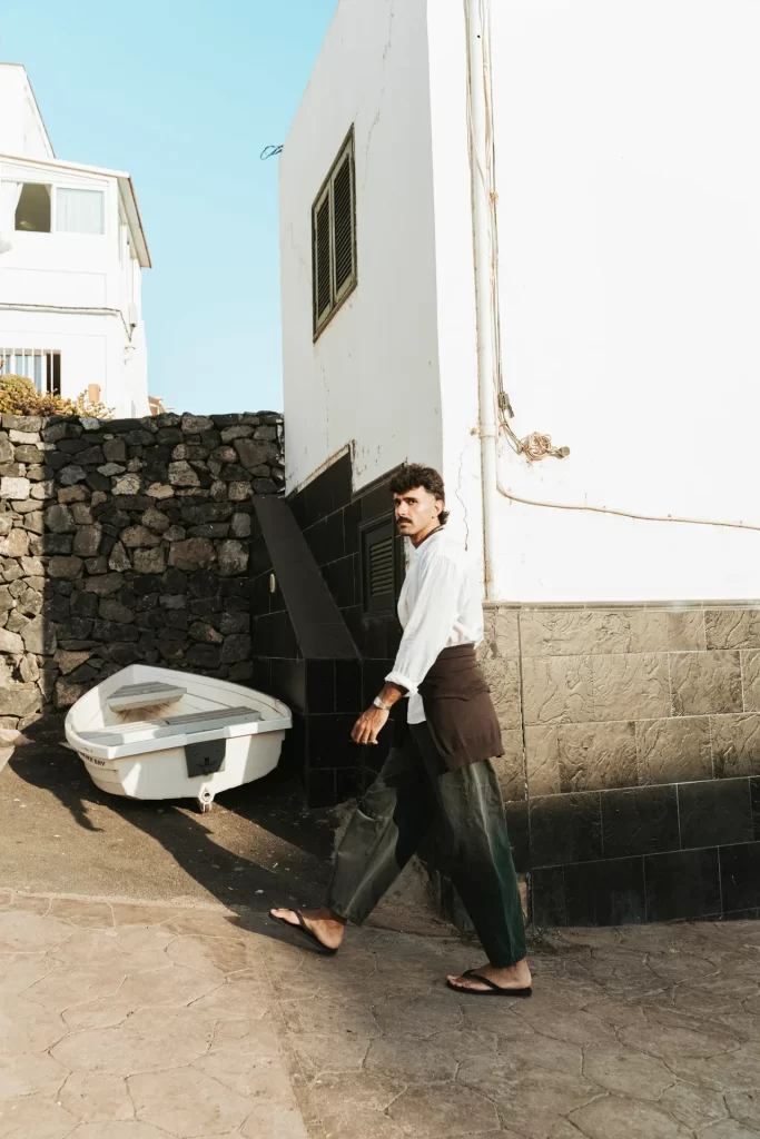 Hombre con bigote caminando por una calle de casas blancas junto a una barca