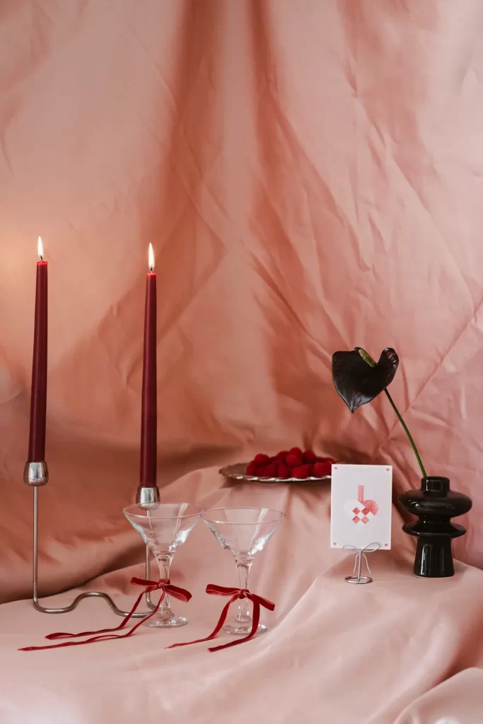 Dos velas granates encendidas, copas con lazos rojos, tarjeta y jarrón negro sobre tela rosa