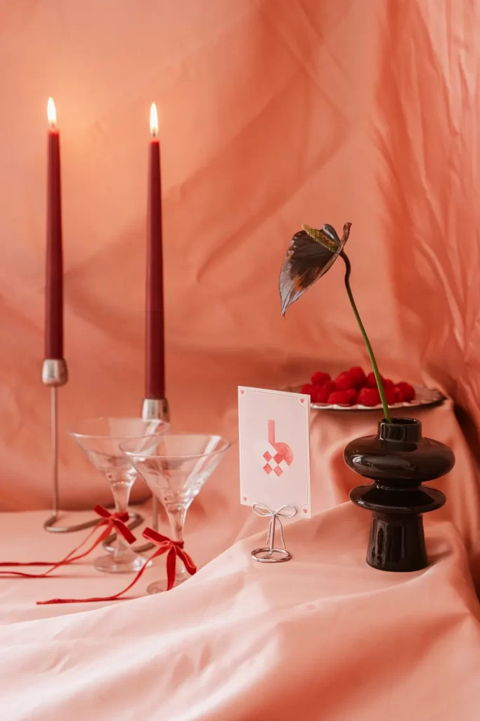 Velas granates encendidas, copas con lazos rojos, tarjeta, frambuesas y jarrón negro sobre tela rosa