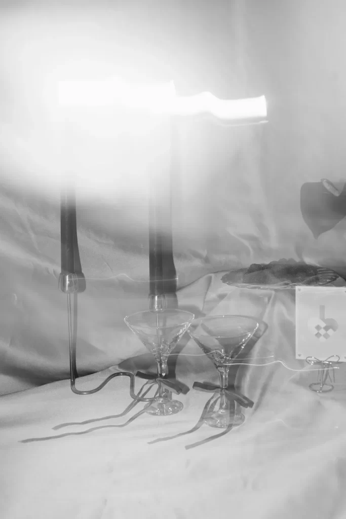 Bodegón en blanco y negro con velas, copas de cóctel con lazos y un destello de luz sobre tela satinada