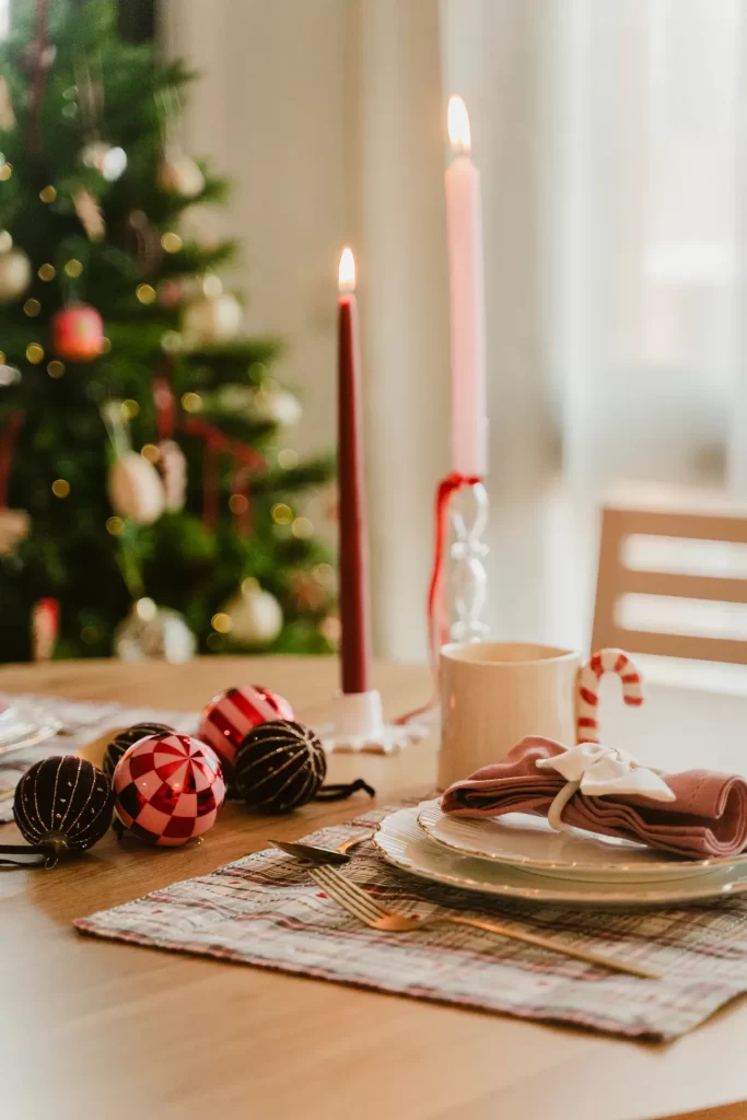 Mesa navideña con velas encendidas, bolas de colores, taza y servilleta rosa ante un árbol de Navidad