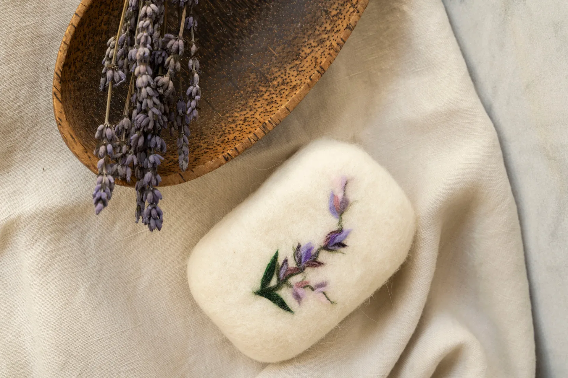 Jabón envuelto en fieltro con una rama de lavanda bordada, junto a un cuenco de madera
