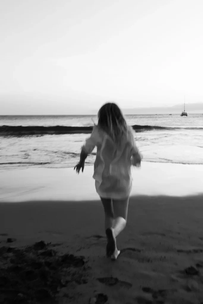 Mujer con camisa blanca corriendo de espaldas hacia la orilla del mar, en blanco y negro