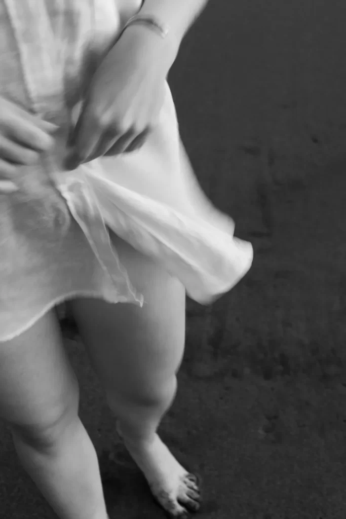 Detalle en movimiento de una camisa blanca agitada y piernas descalzas sobre arena oscura, en blanco y negro