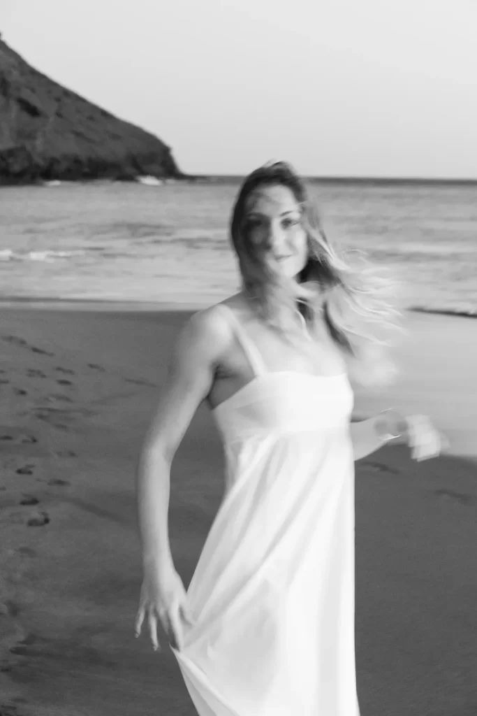 Mujer con vestido blanco en movimiento en la orilla de una playa, en blanco y negro