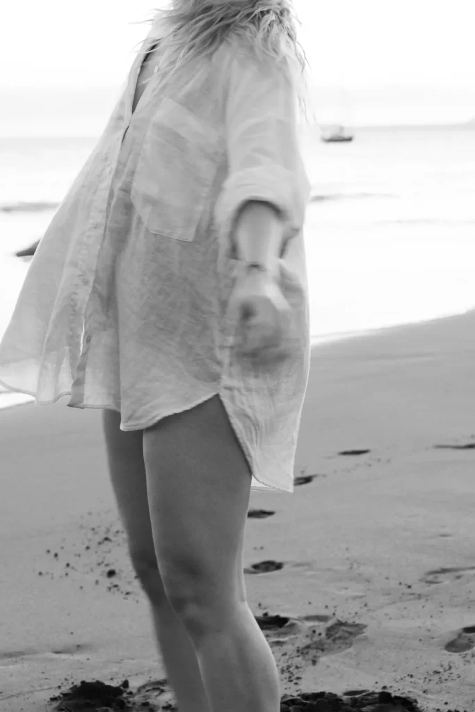 Mujer con camisa amplia de lino en la playa con huellas en la arena y un velero al fondo