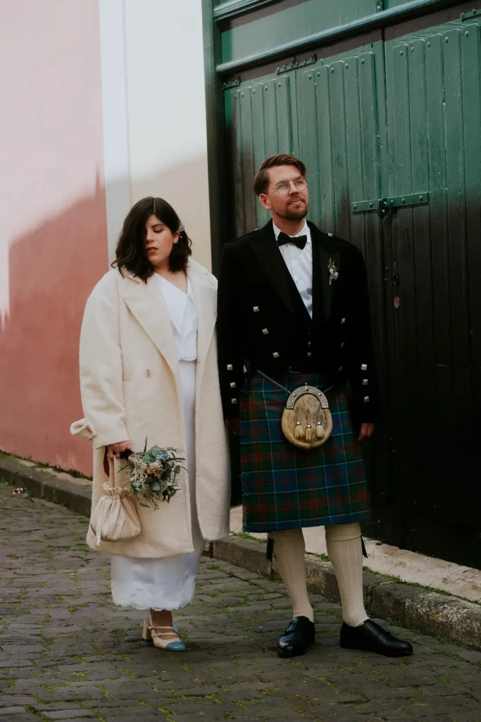 Novia con abrigo blanco y ramo junto al novio con kilt ante un portón verde en una calle empedrada
