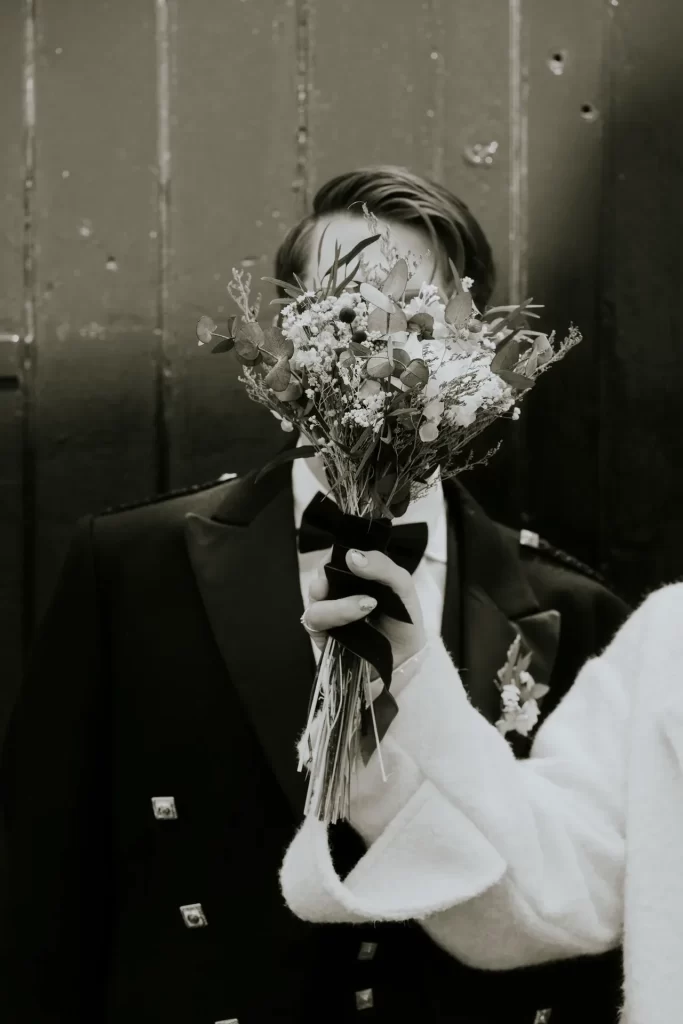 Novio con pajarita tapado por un ramo de flores secas que sostiene la mano de la novia