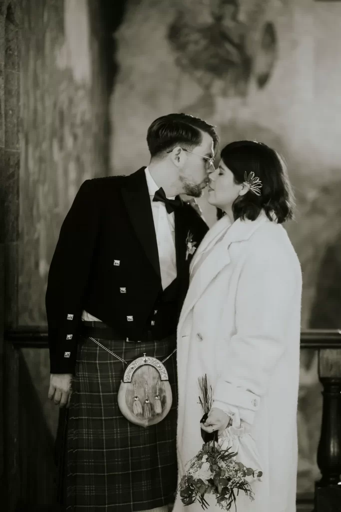 Novio con kilt y novia con abrigo blanco rozándose la nariz ante un mural pintado