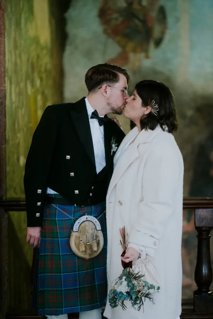 Novios besándose ante un mural, él con kilt de tartán y ella con abrigo blanco y ramo