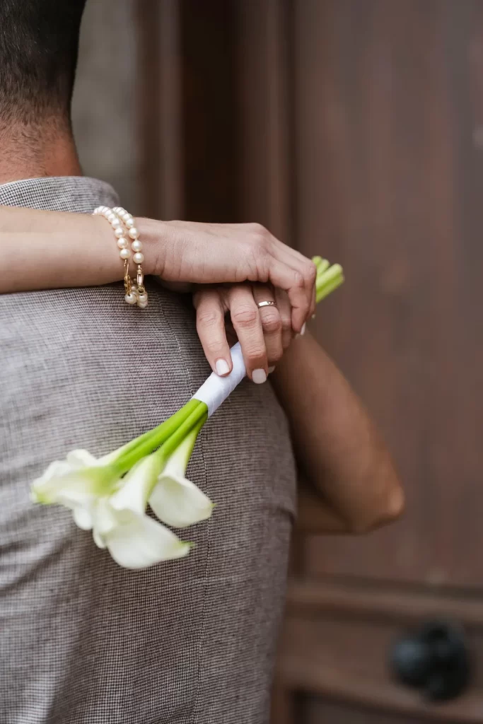 Novia abrazando al novio por la espalda con un ramo de calas blancas y pulsera de perlas