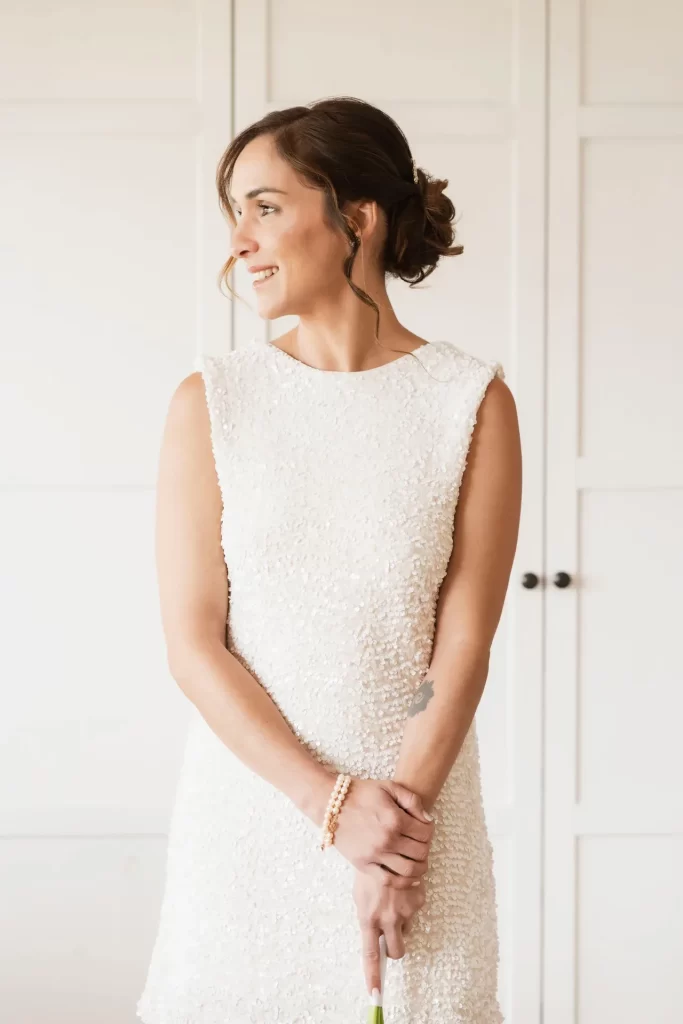 Novia sonriente con vestido corto de lentejuelas mirando de lado ante un armario blanco