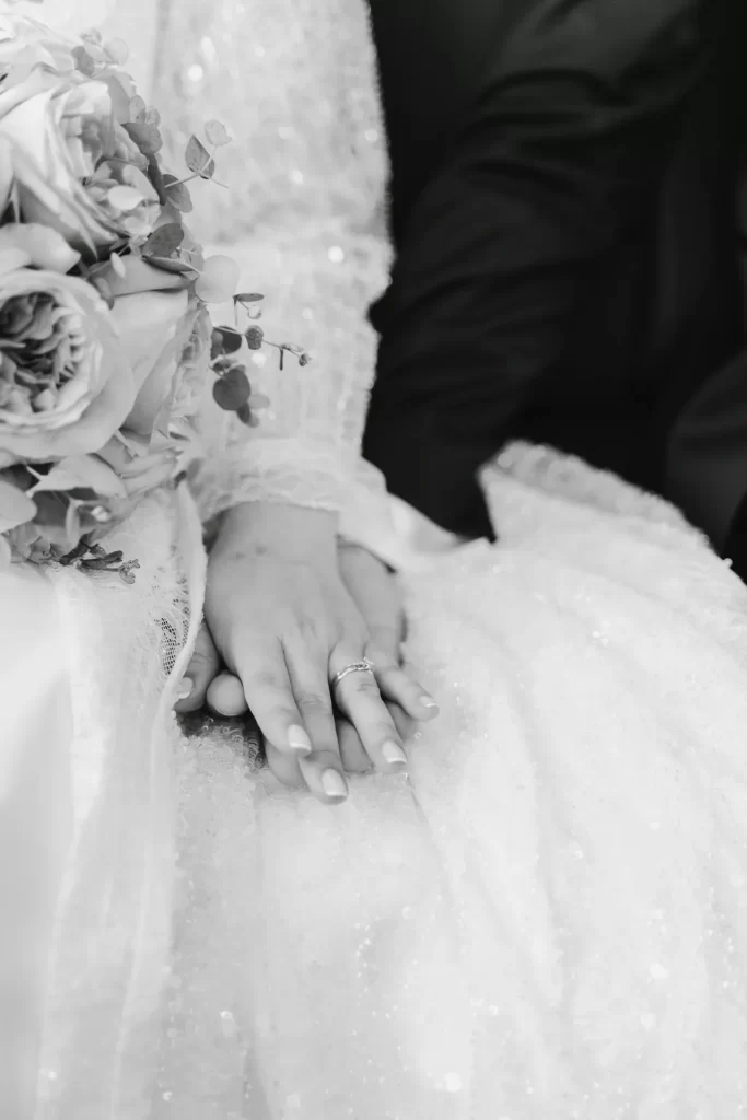 Mano de la novia con anillo sobre la del novio, apoyadas en el vestido junto al ramo