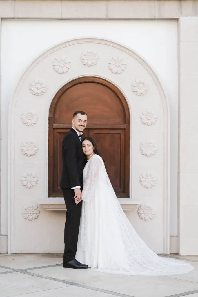 Novios abrazados ante una puerta de madera con arco blanco decorado con rosetones