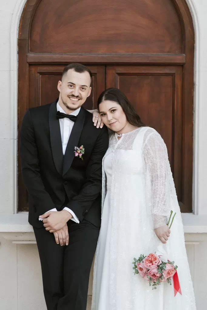 Novios sonrientes apoyados ante un portón de madera, ella con ramo de rosas y cinta roja