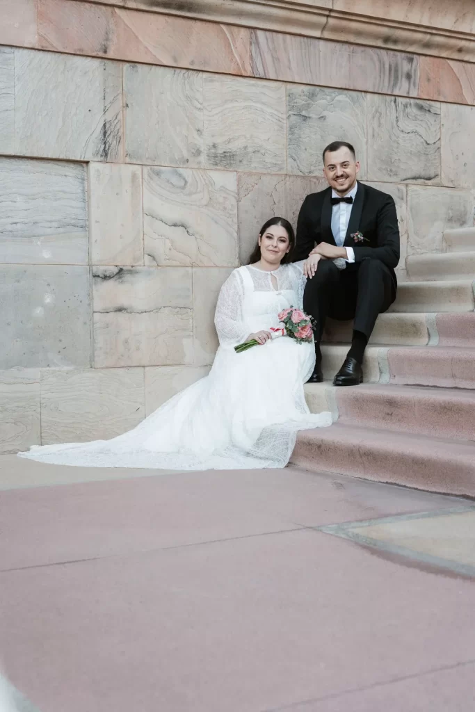 Novios sentados en una escalinata de piedra junto a un muro de sillares, ella con ramo